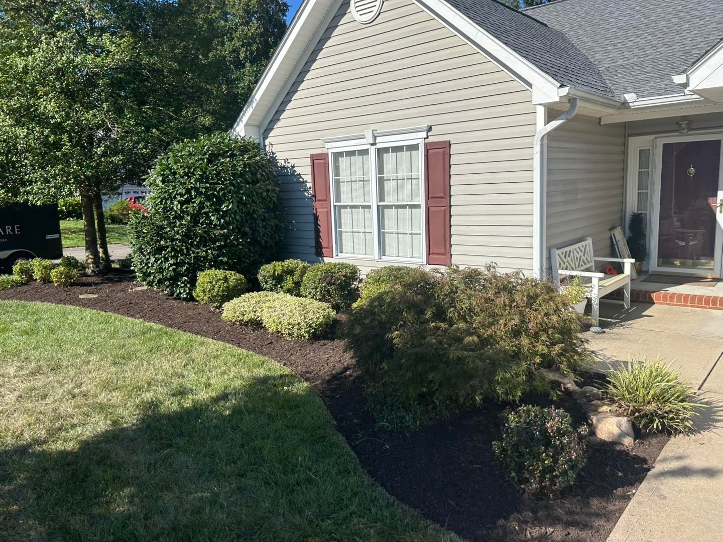 A tan-sided house with a dark door and windows with maroon shutters, fronted by a mulched garden bed with shrubs and a bench.
