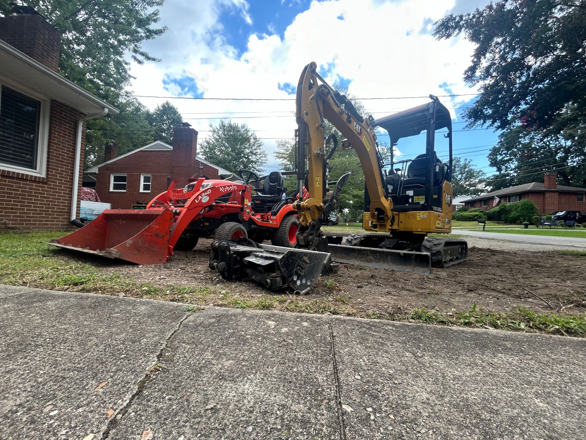 A yellow mini excavator and a red tractor parked on a grassy residential lot next to a brick house.
