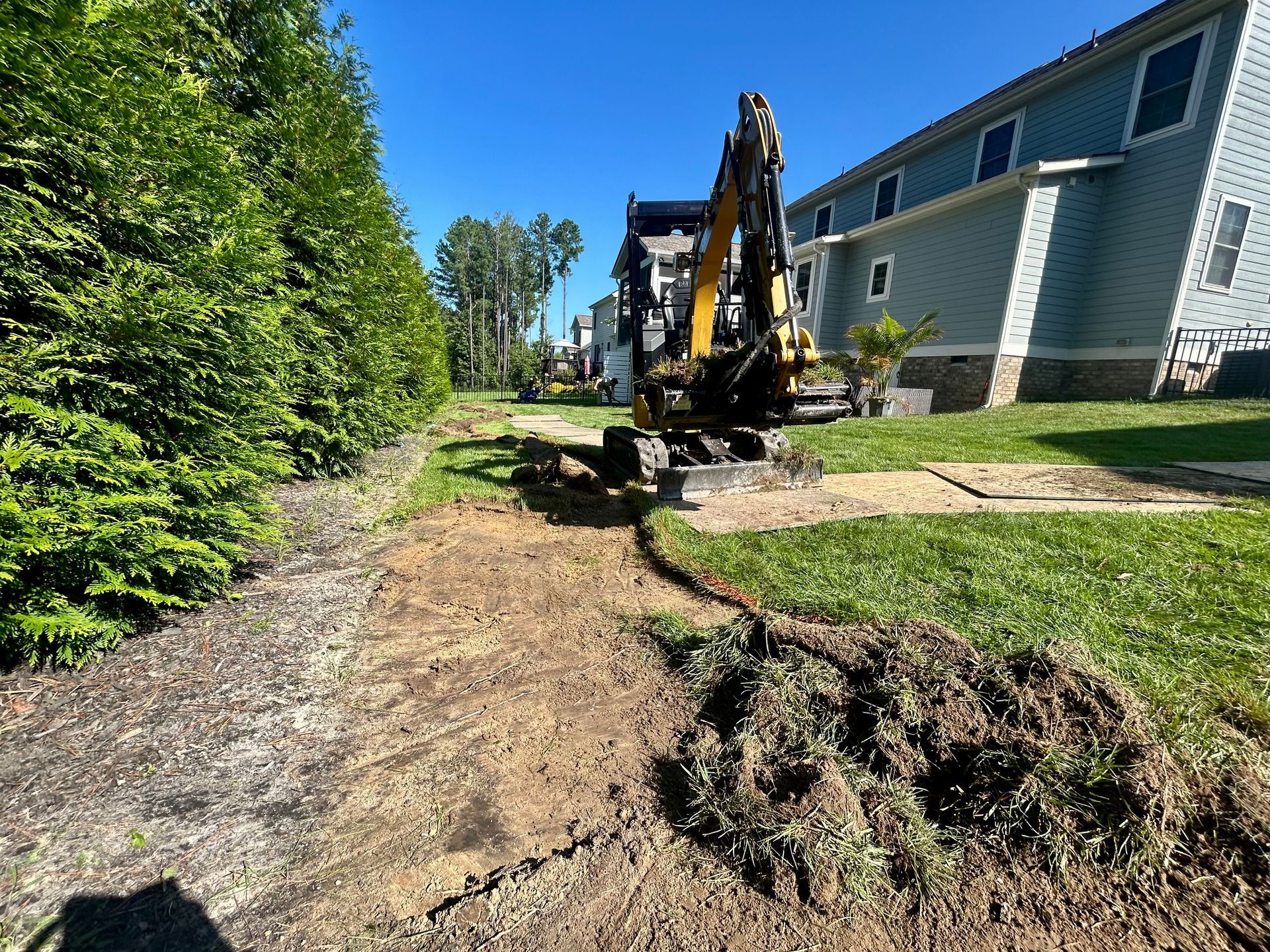 A yellow mini excavator sits on a dirt path next to a residential house and a tall hedge on a sunny day.