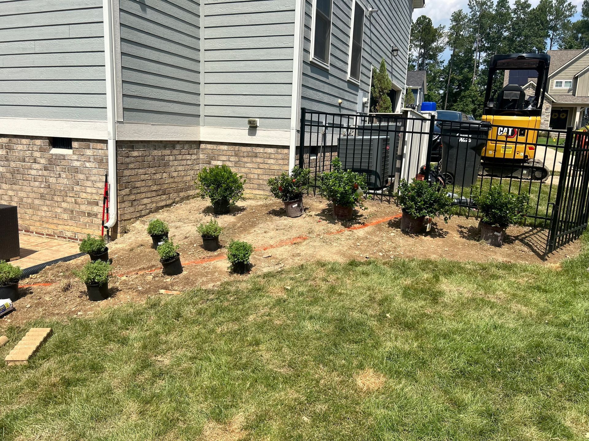 Newly planted bushes in a mulched garden bed along the side of a house with stone siding and a yellow excavator nearby.
