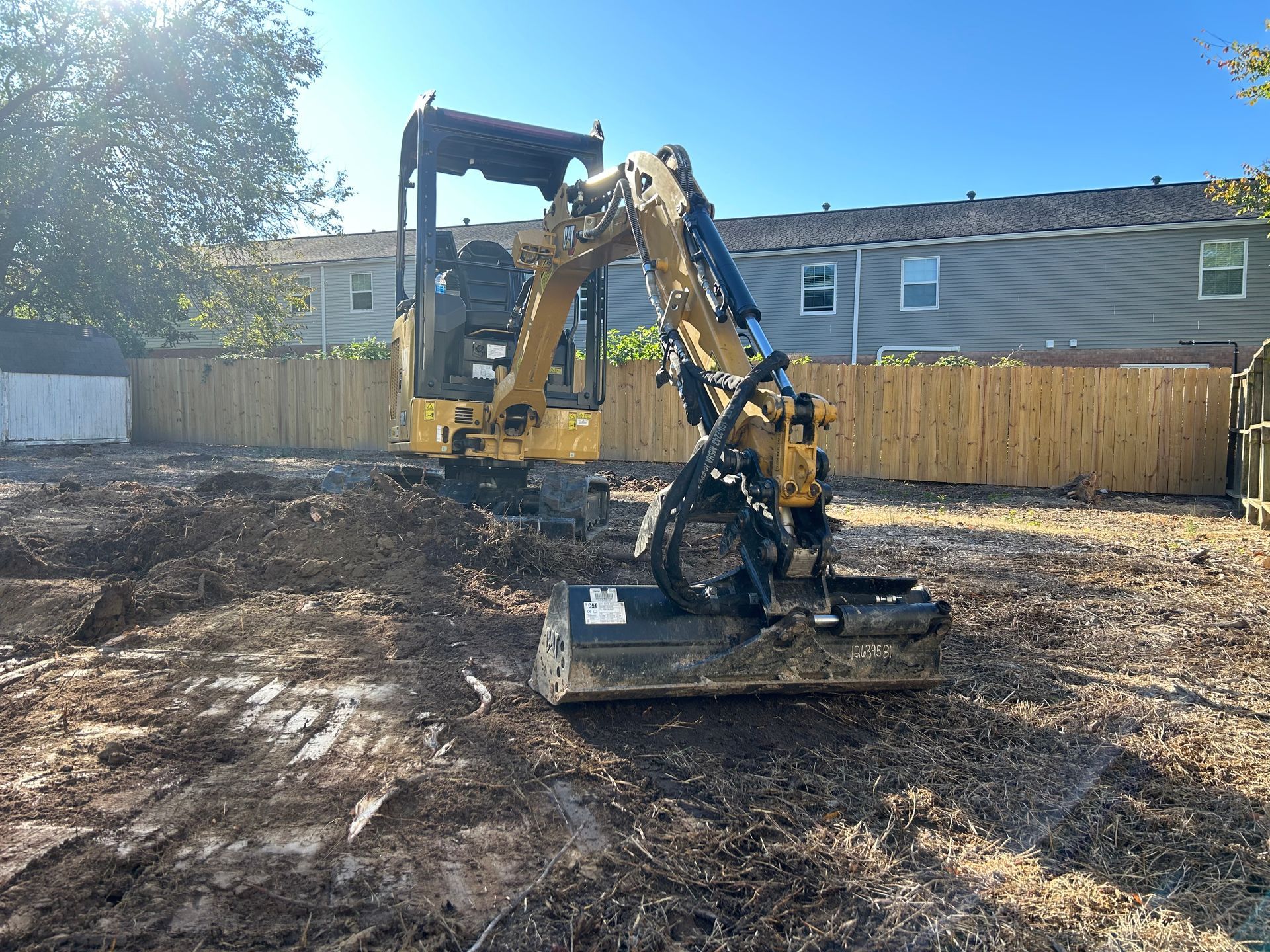 A yellow Caterpillar mini excavator with a mulching attachment parked on dirt in a residential backyard.