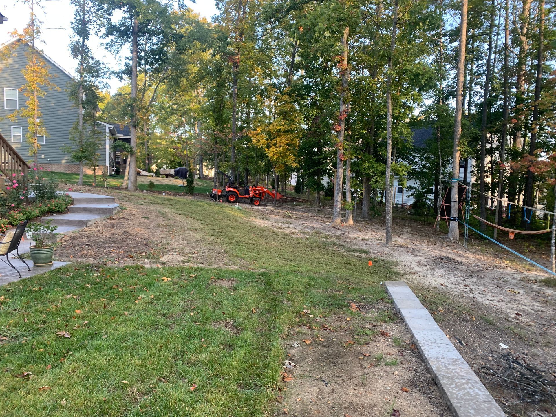 A residential backyard with a grassy lawn, a stone walkway, an orange tractor, and trees in the background.