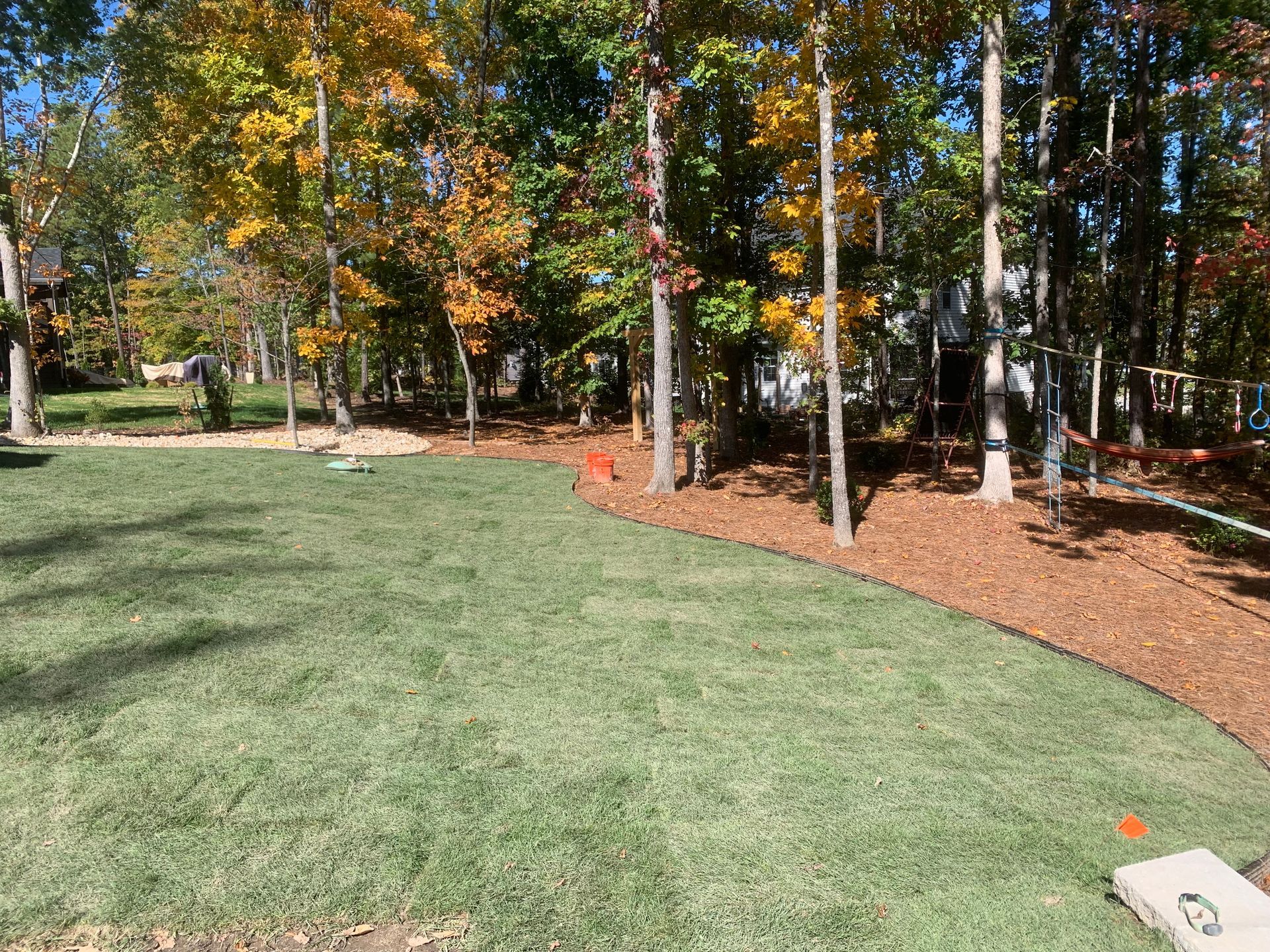 A freshly laid lawn transitions into a curved mulched bed beneath a line of trees with autumn foliage.