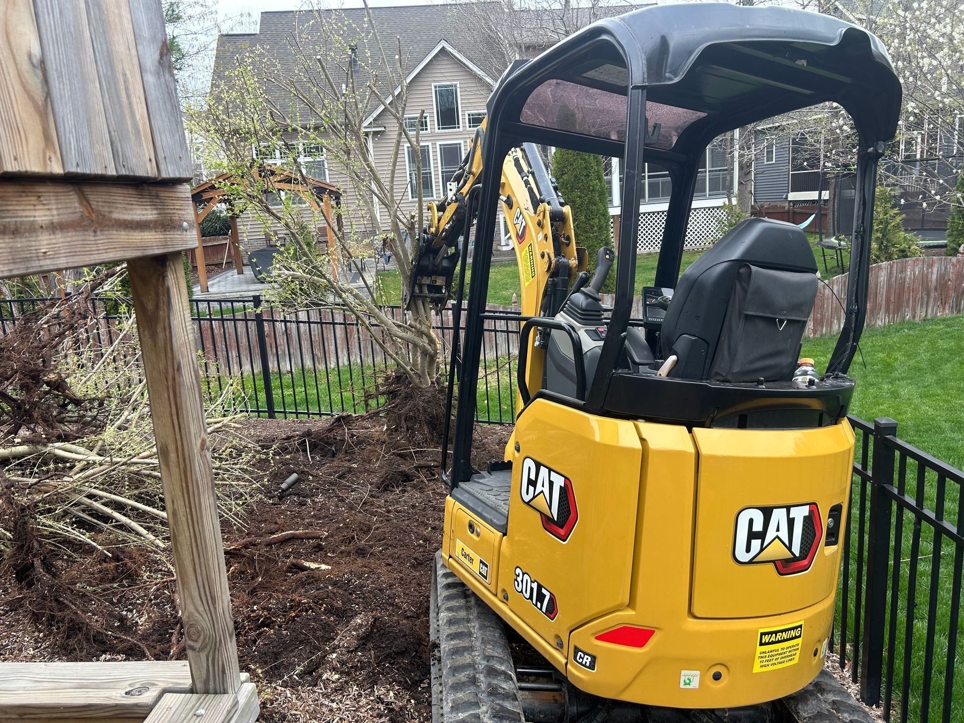 A yellow CAT compact excavator sits in a grassy backyard near a wooden structure, working on clearing a small tree.
