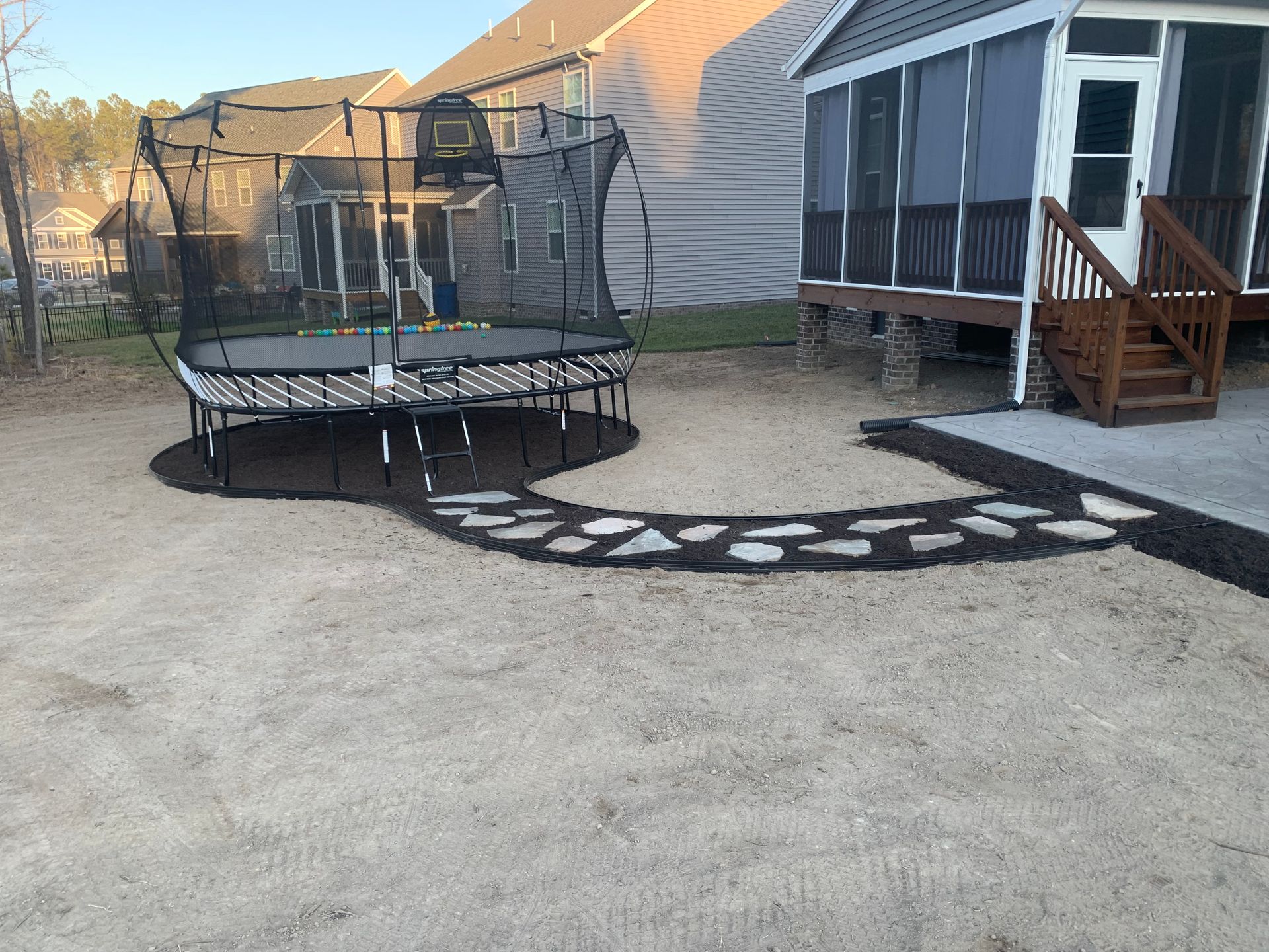 A backyard with a trampoline on mulch, connected by a stone path to a screened-in porch with wooden stairs.