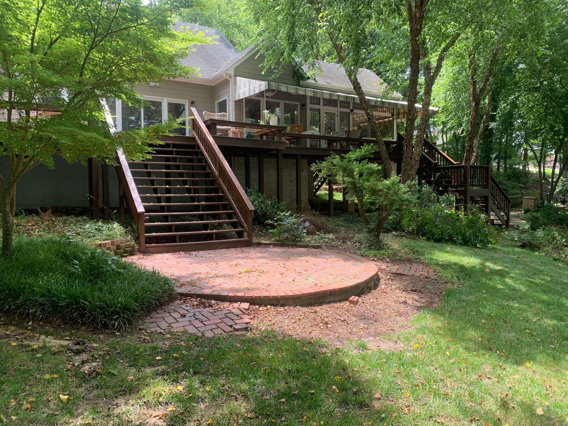 A raised wooden deck with a staircase leading to a circular brick patio in a leafy, shaded backyard.