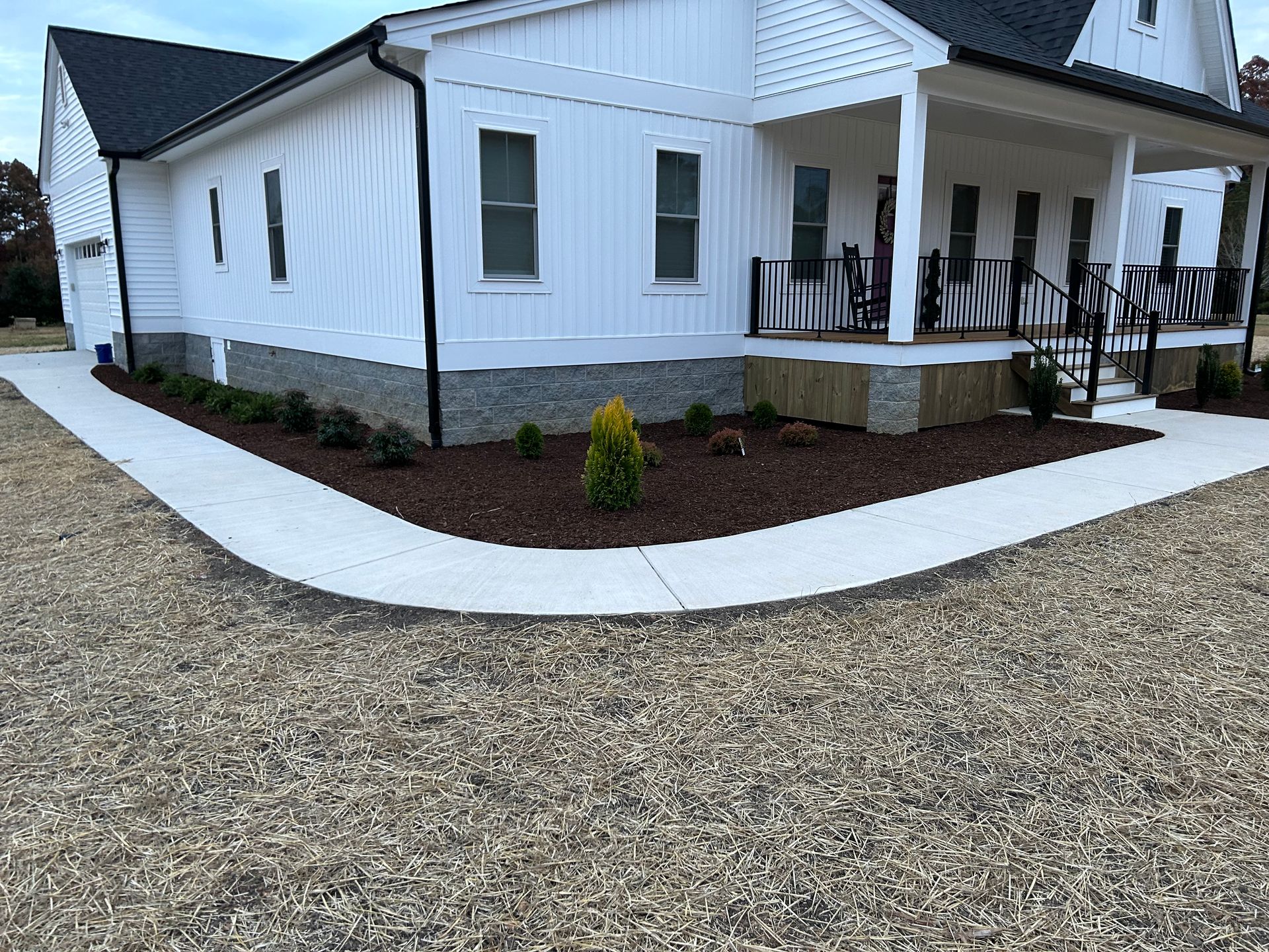 A white farmhouse with a stone foundation and a covered porch, bordered by a concrete walkway and gravel driveway.