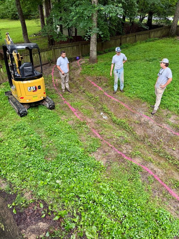 A backyard garden bed with mulch, small shrubs, and a tree, located next to a gray house with a black fence in the distance.