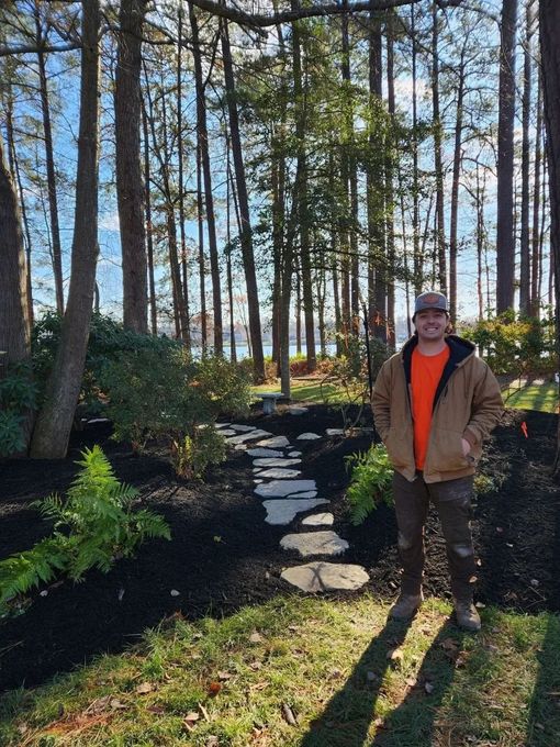 A stone-edged flower bed with mulch and small green and yellow shrubs next to a concrete sidewalk and a house porch.