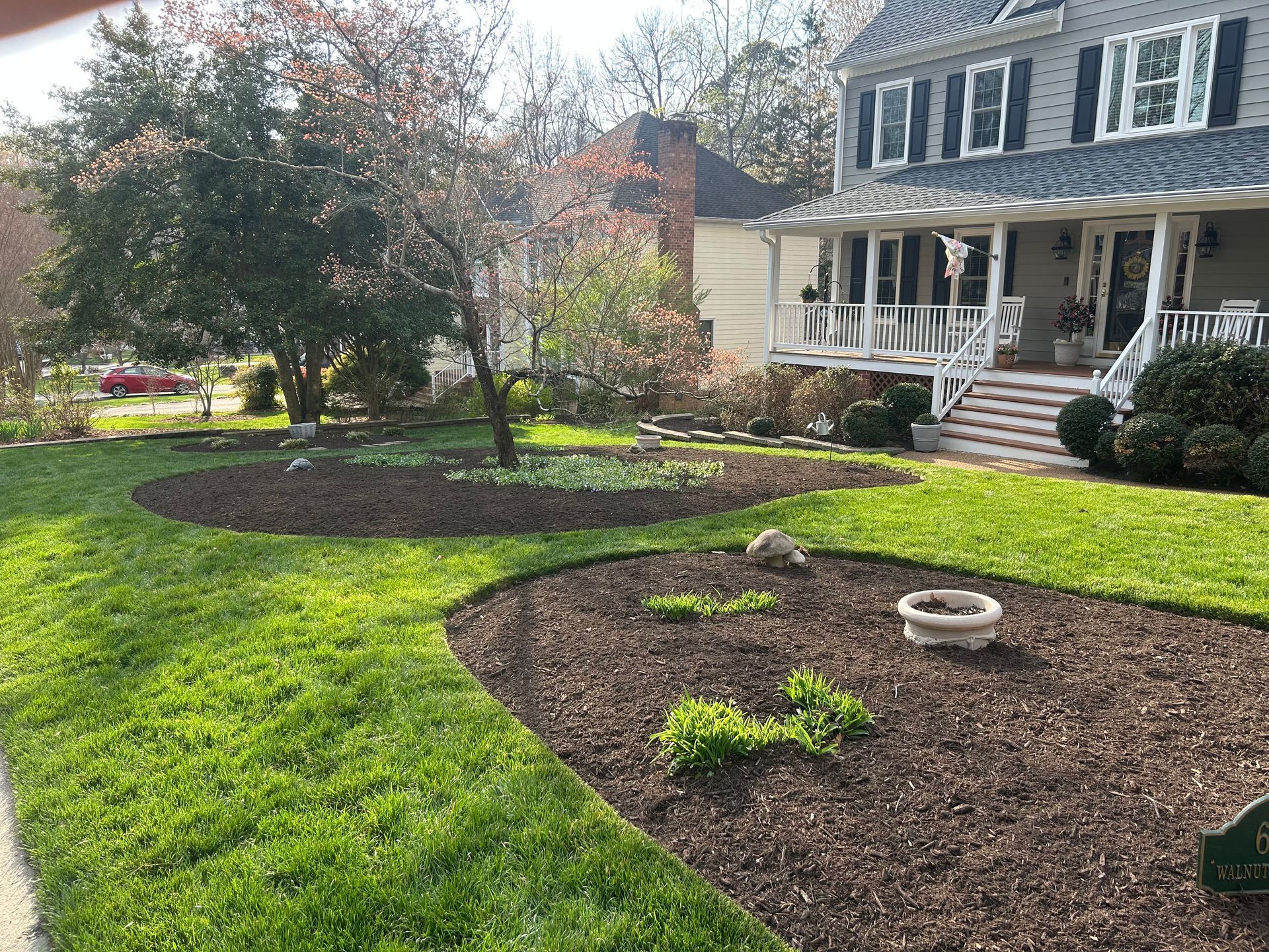 A white, single-story house exterior features a manicured green lawn, mulch landscaping, and trees against a blue sky.