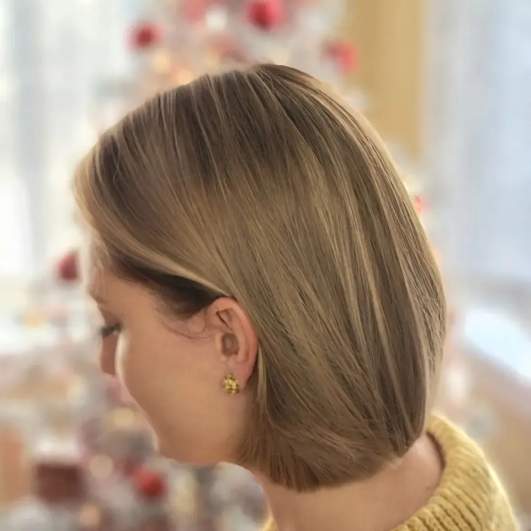 A woman wearing a yellow sweater and earrings is standing in front of a christmas tree.