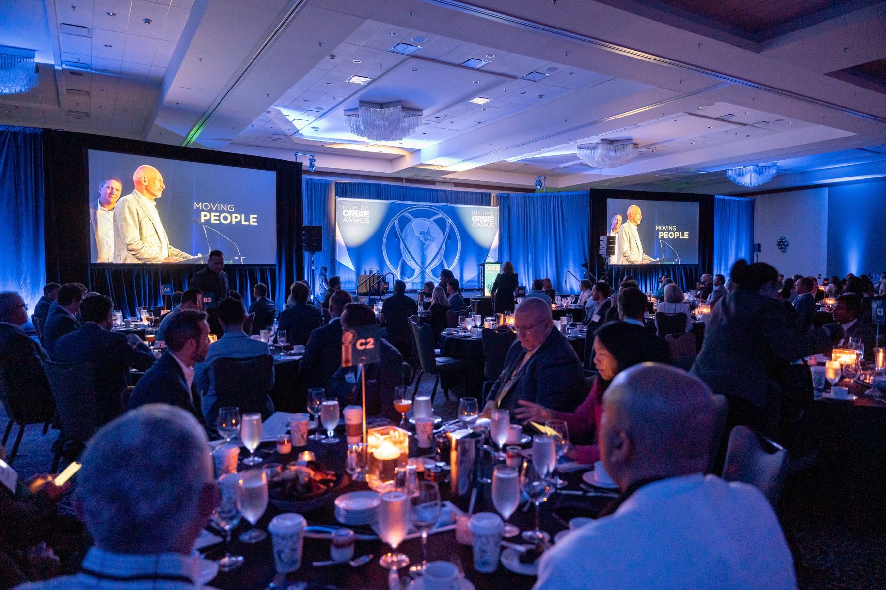 A large group of people are sitting at tables in a large room watching a presentation.