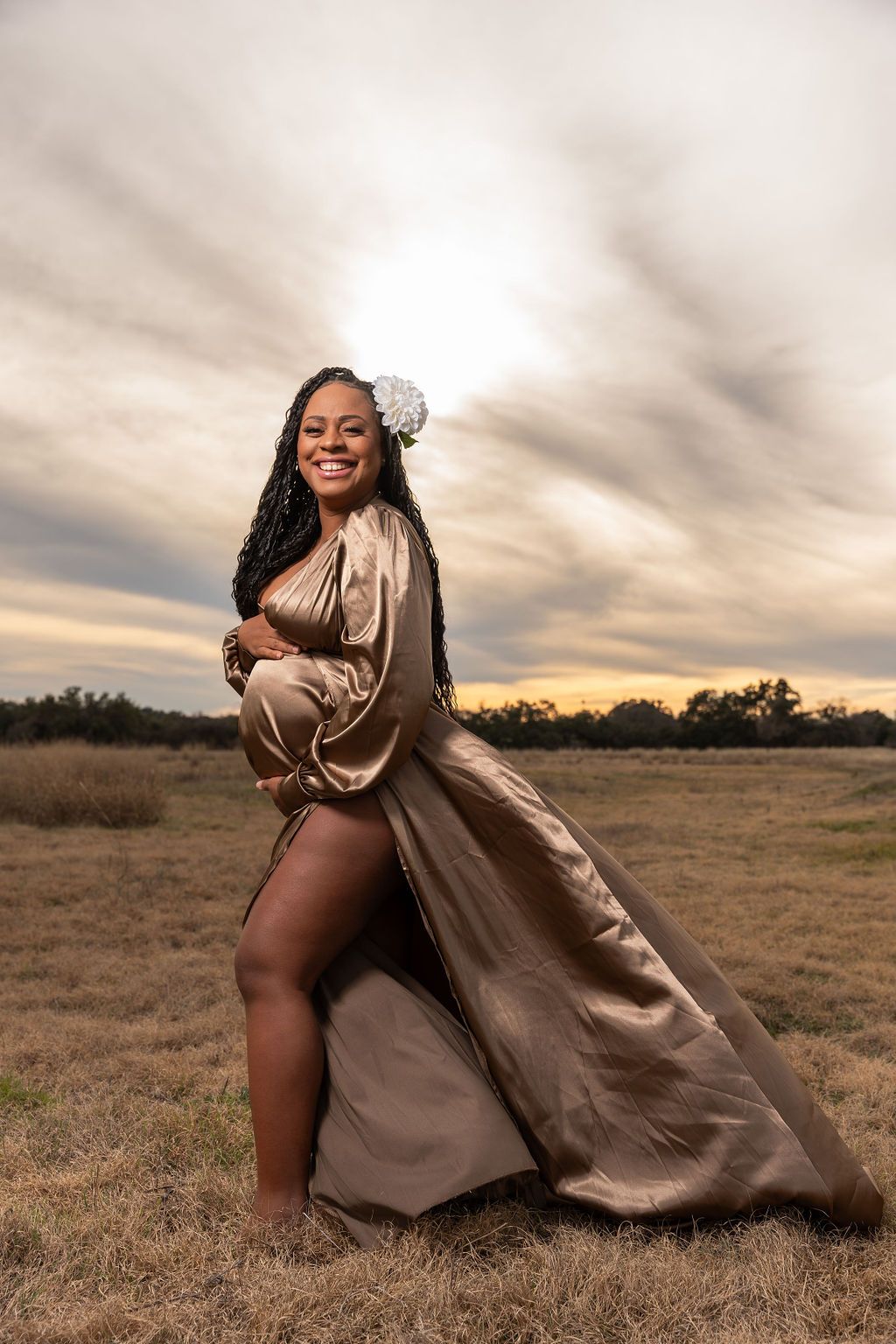 A pregnant woman in a long gold dress is kneeling in a field.