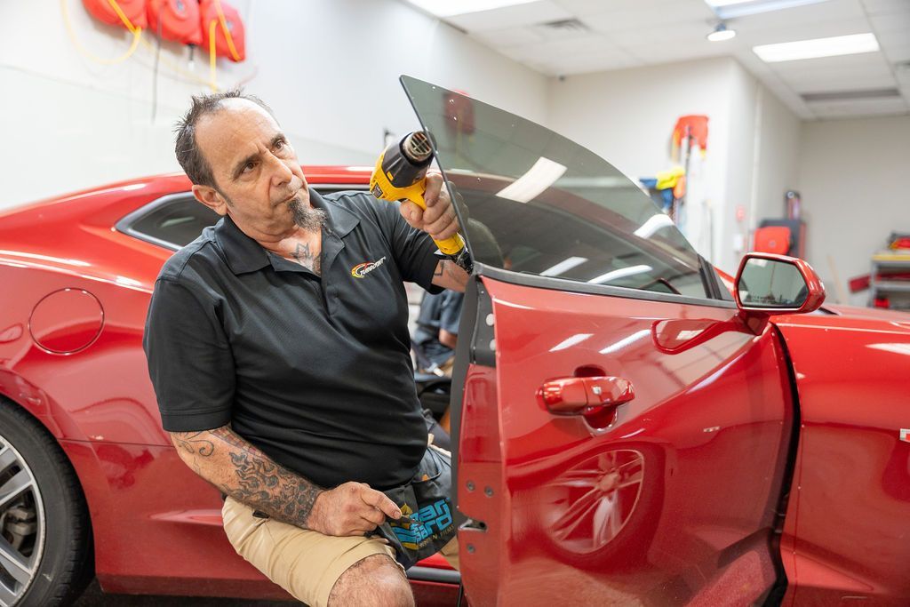 A man is sitting on the side of a red car holding a tool.