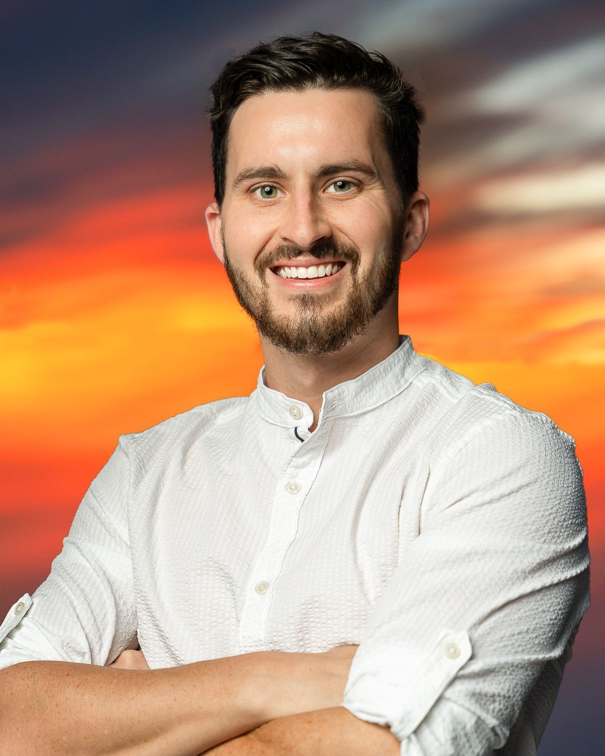 A man with a beard is smiling with his arms crossed in front of a sunset.