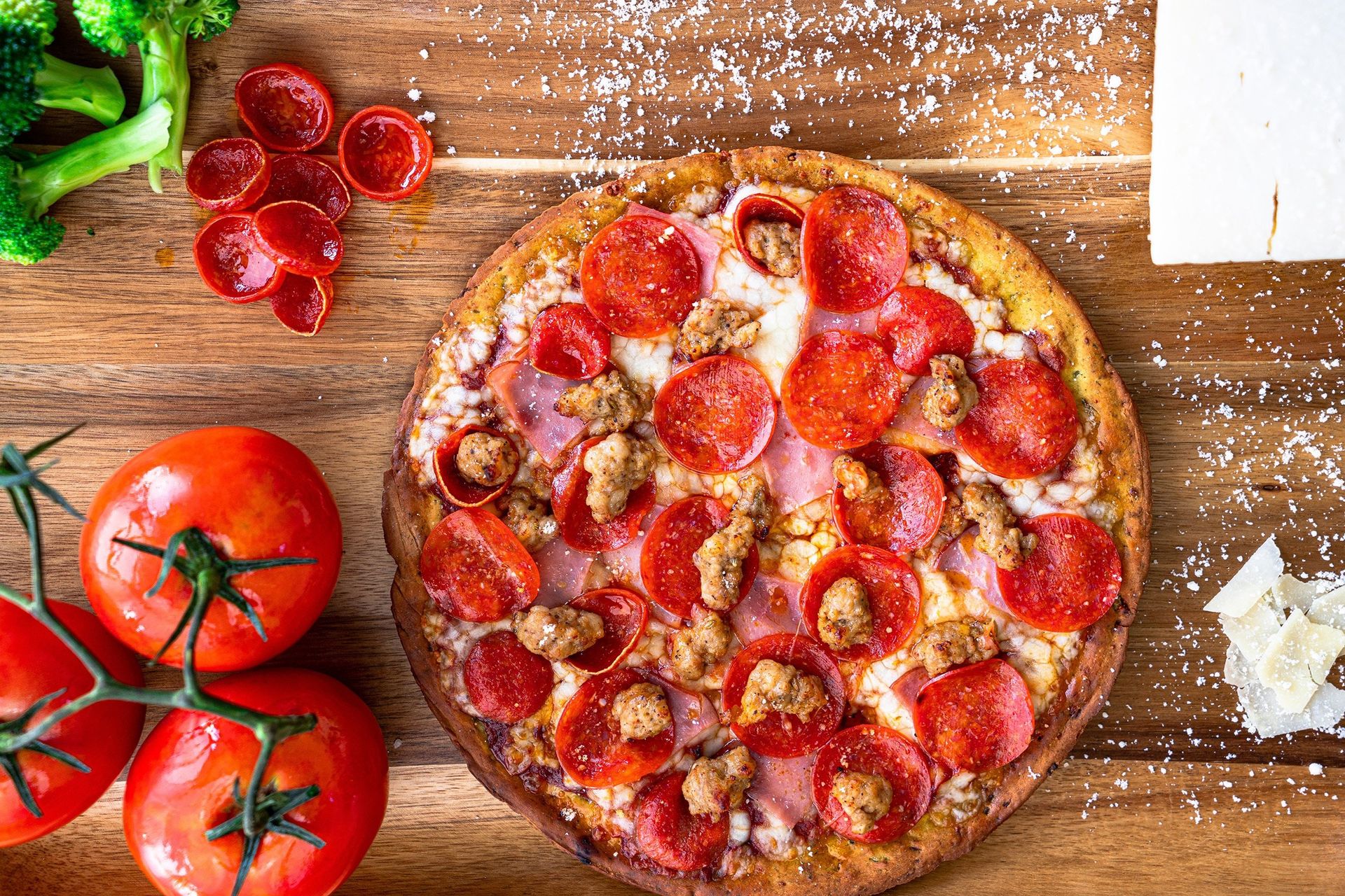 A pepperoni pizza is sitting on a wooden table next to tomatoes and broccoli.