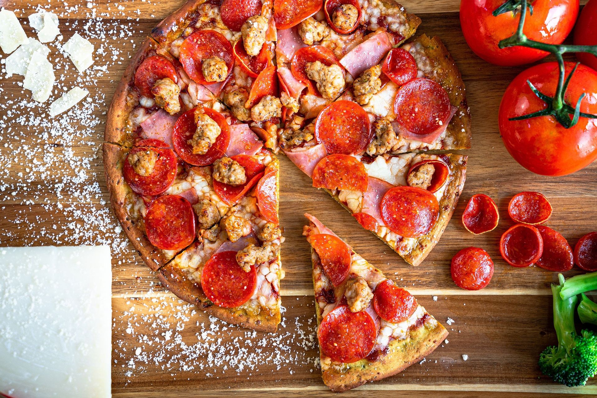 A pepperoni pizza with tomatoes and broccoli on a wooden cutting board.