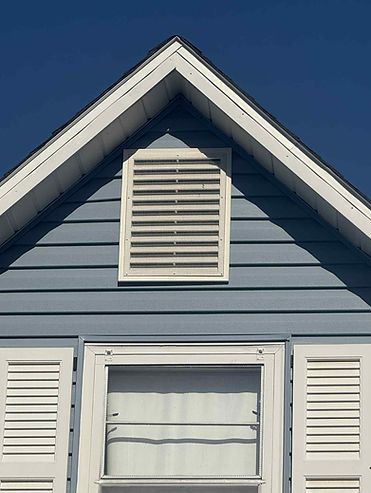 Blue house gable with white trim, vent, and window with shutters against a clear sky.