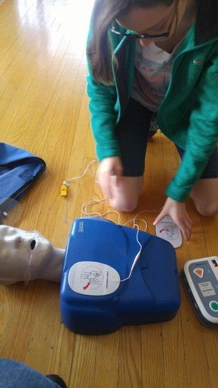 A woman is kneeling on the floor using a defibrillator on a mannequin.
