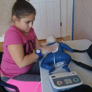 A young girl is sitting at a table using a defibrillator on a mannequin.