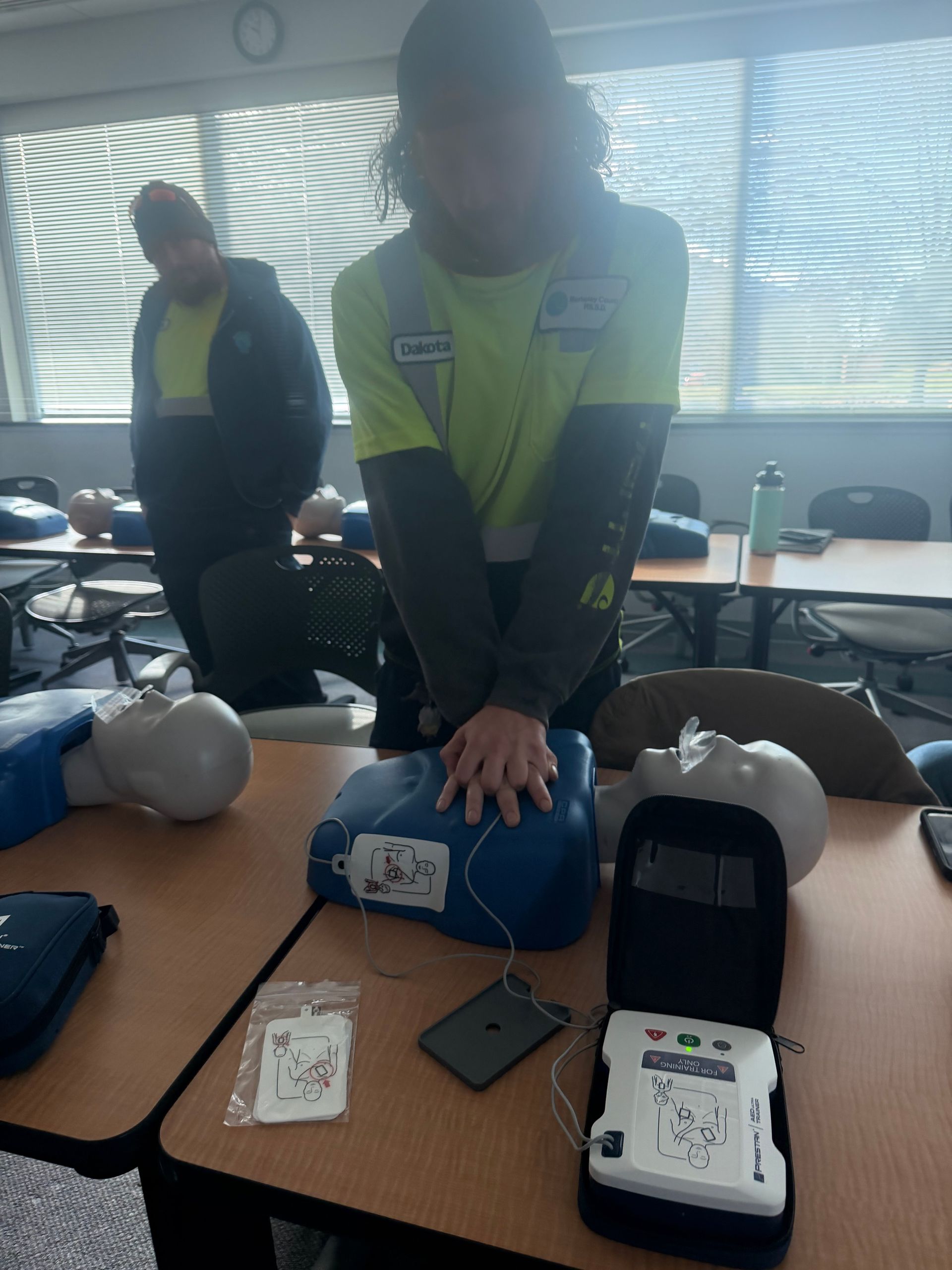 Person demonstrating CPR on a mannequin in a classroom, with another person observing. An AED is open on the table.