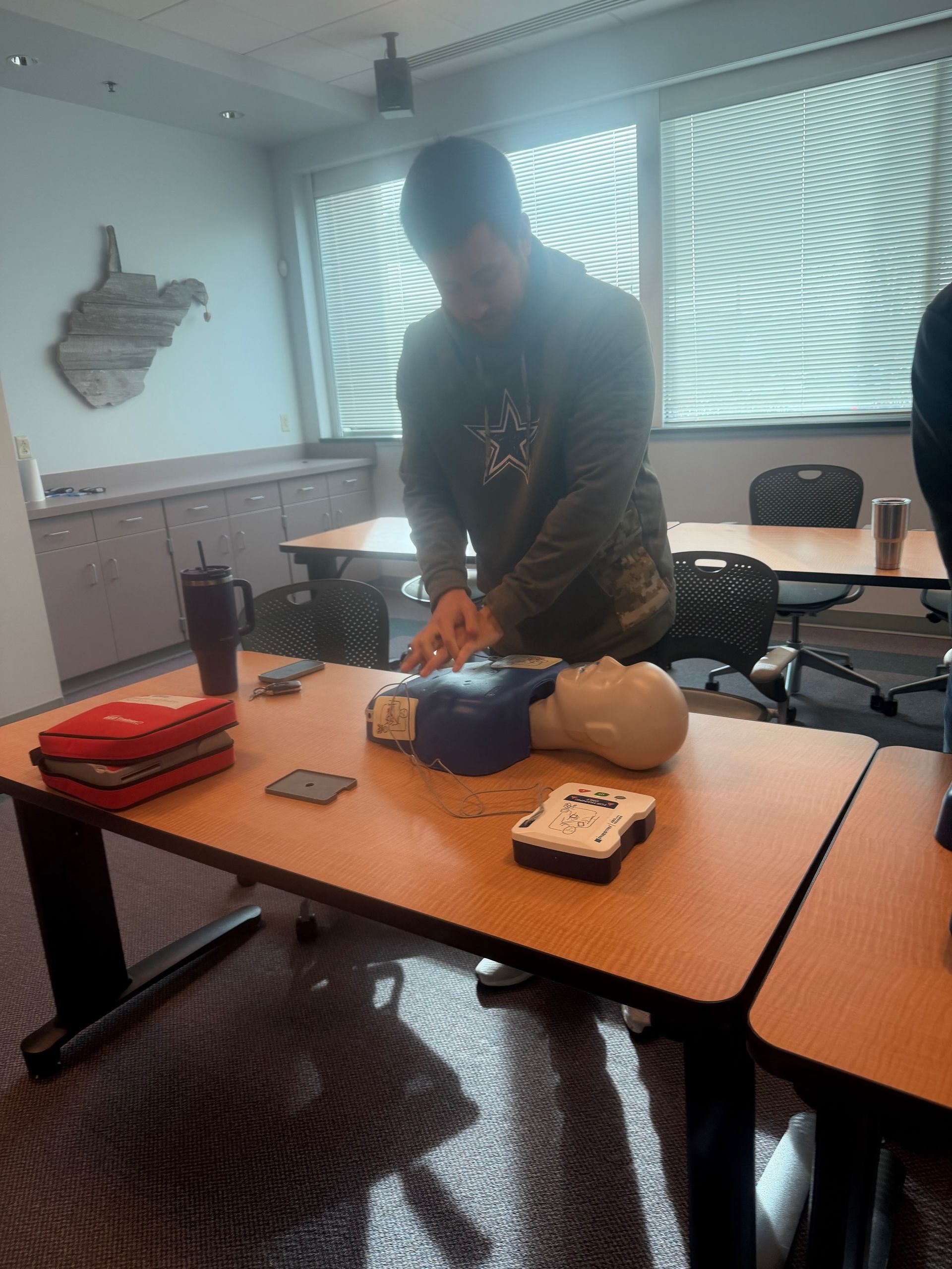 Person practicing CPR on a training dummy, using an AED. Classroom setting with windows and a map of West Virginia.