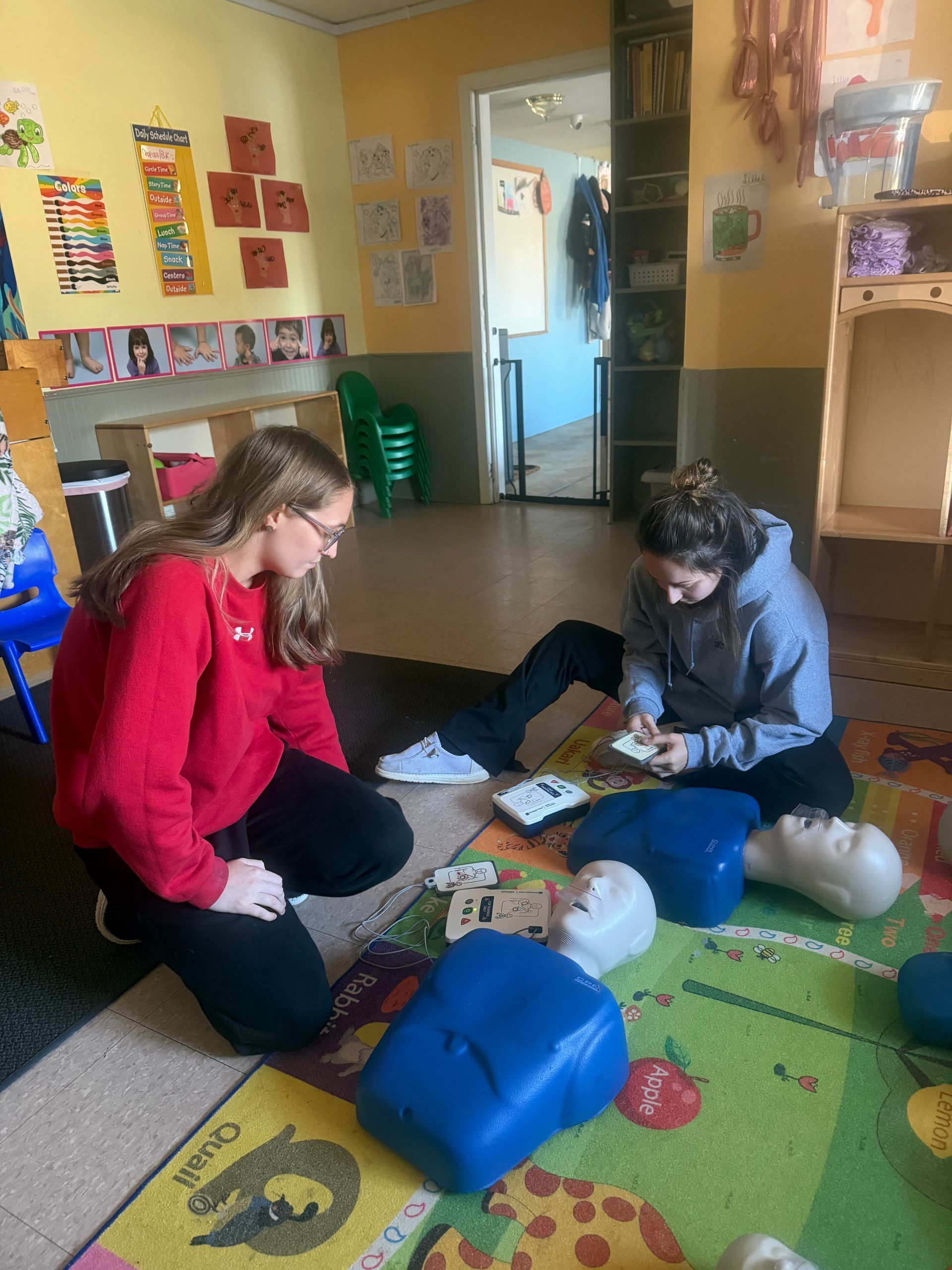 Two people kneeling on a rug, practicing CPR on mannequins in a classroom.