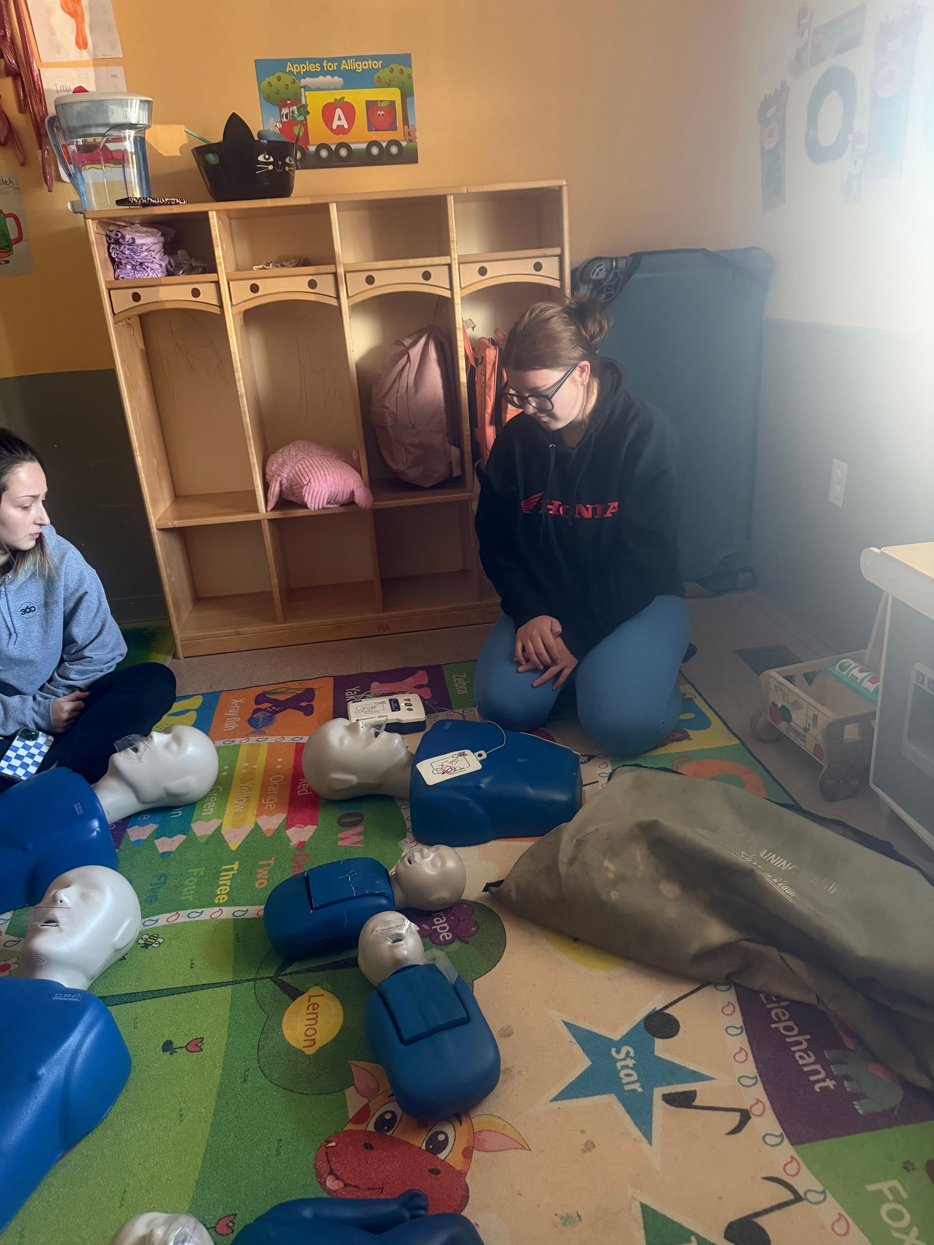 Woman kneeling, demonstrating CPR on a training dummy, surrounded by other dummies. Indoor setting.