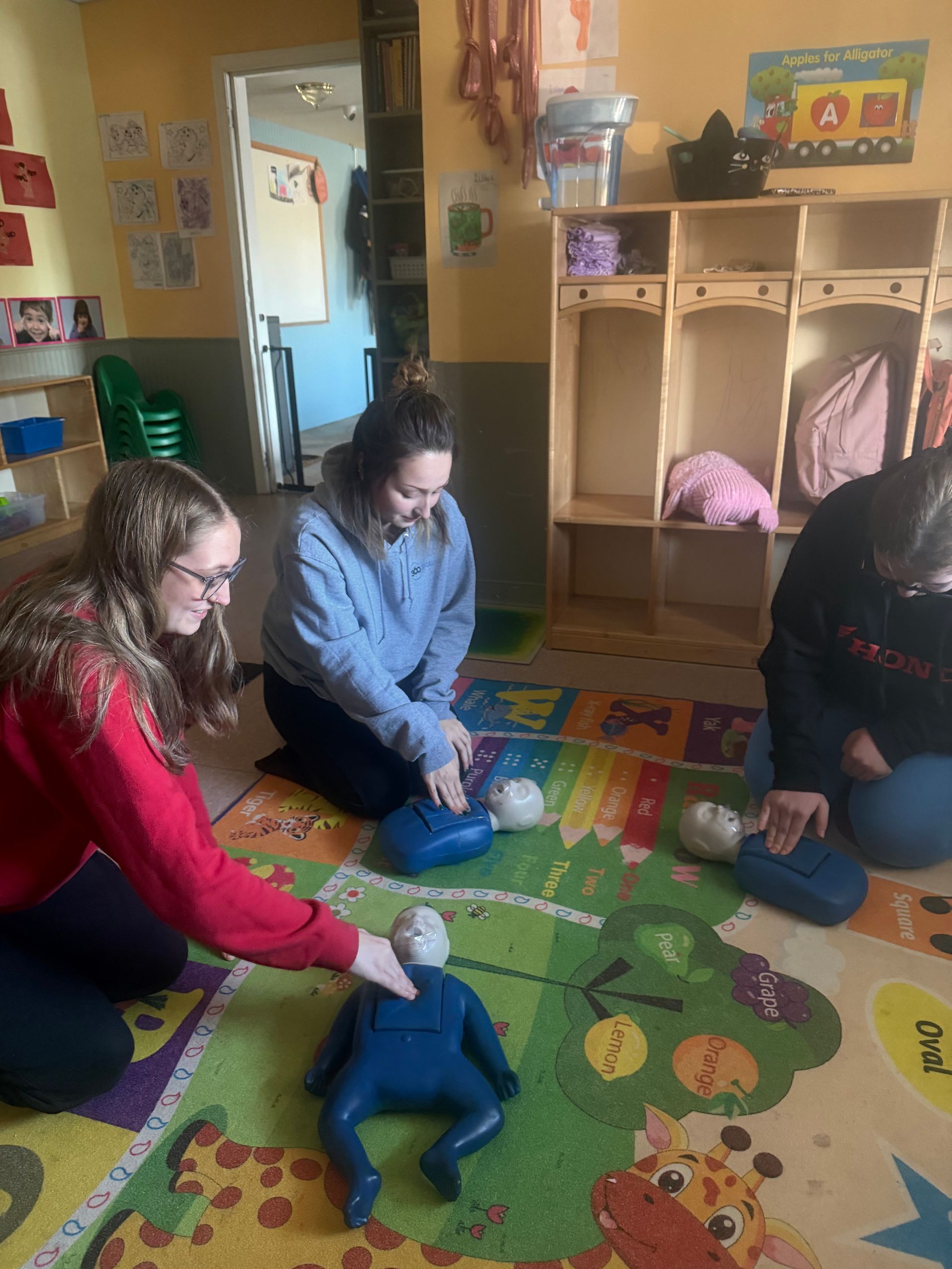 Three people practice CPR on child-sized dummies in a classroom, one person points.