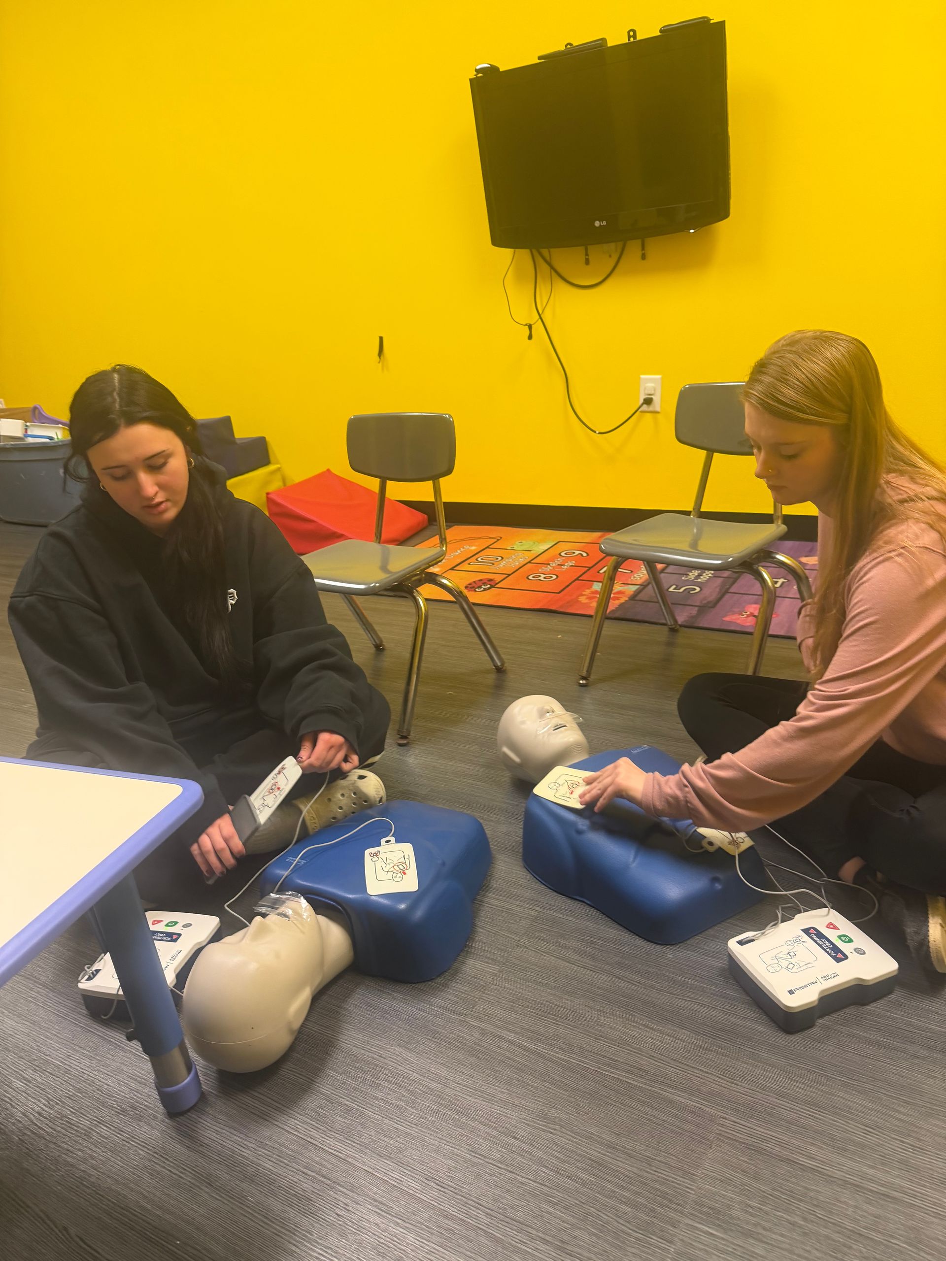 Two people practicing CPR on mannequins with AEDs in a room with yellow walls.