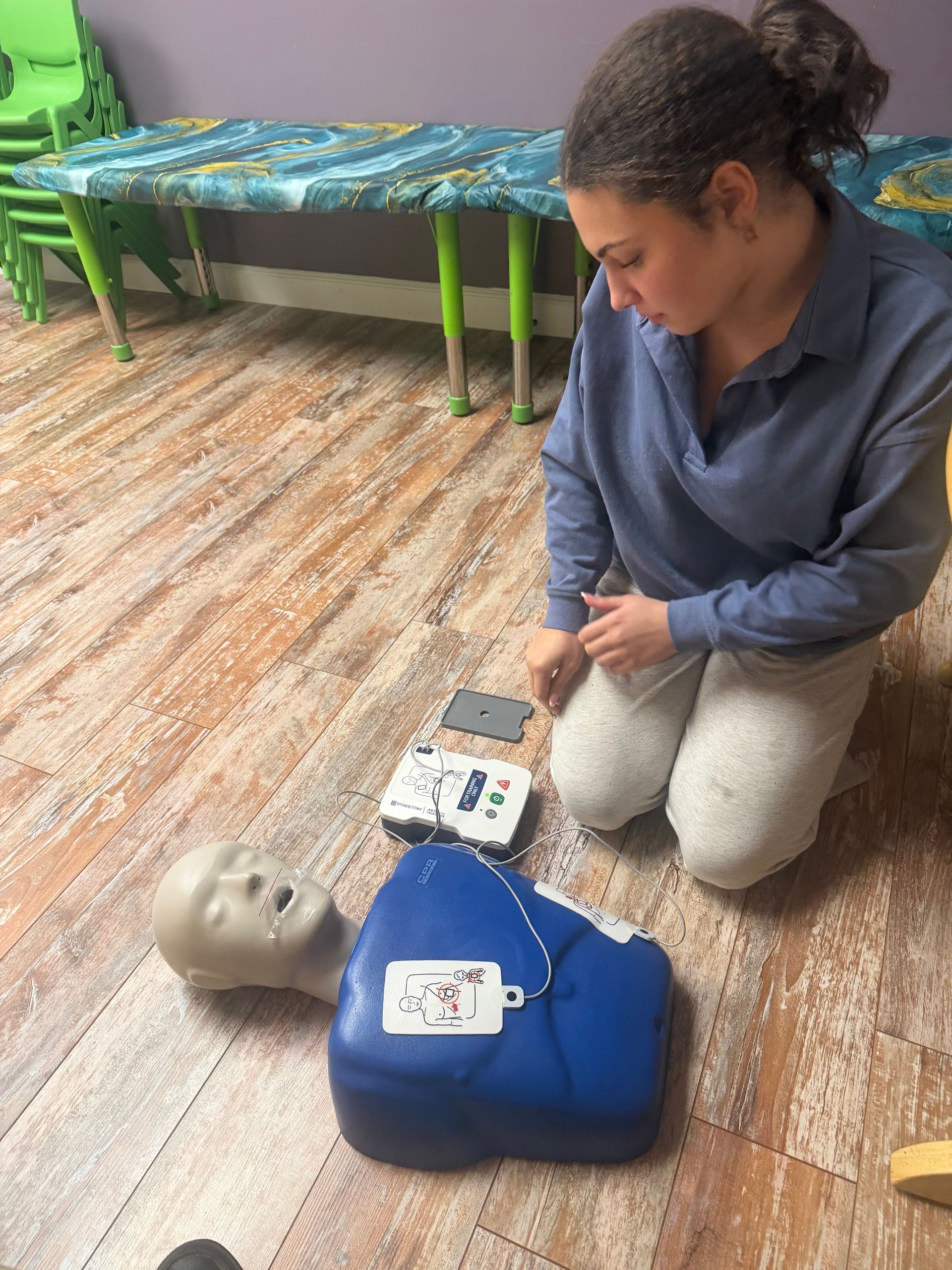 Woman kneeling near a CPR dummy, demonstrating AED use on a wooden floor in a room.