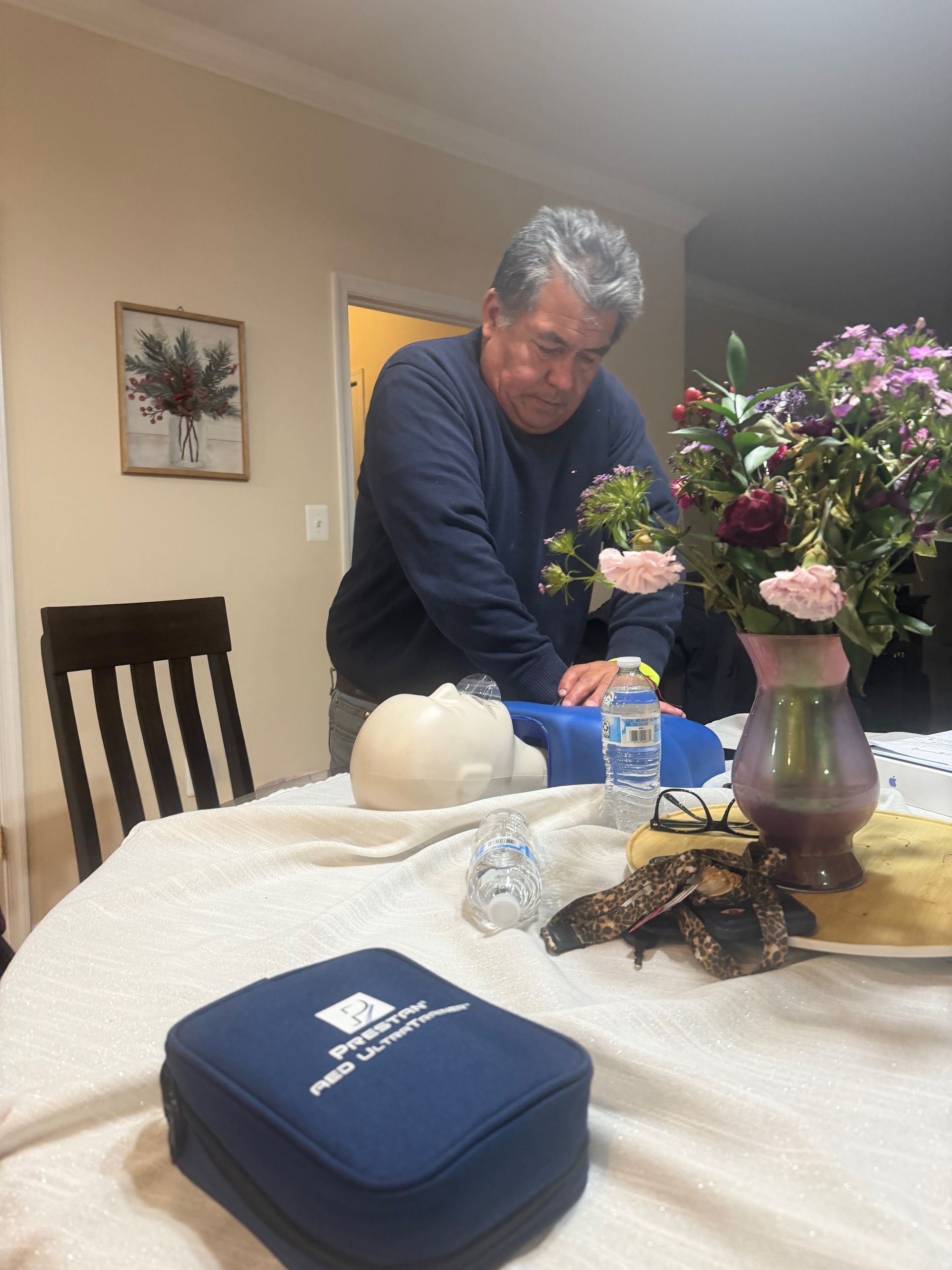 Man practicing CPR on a training dummy, indoors. Table with kit, flowers.