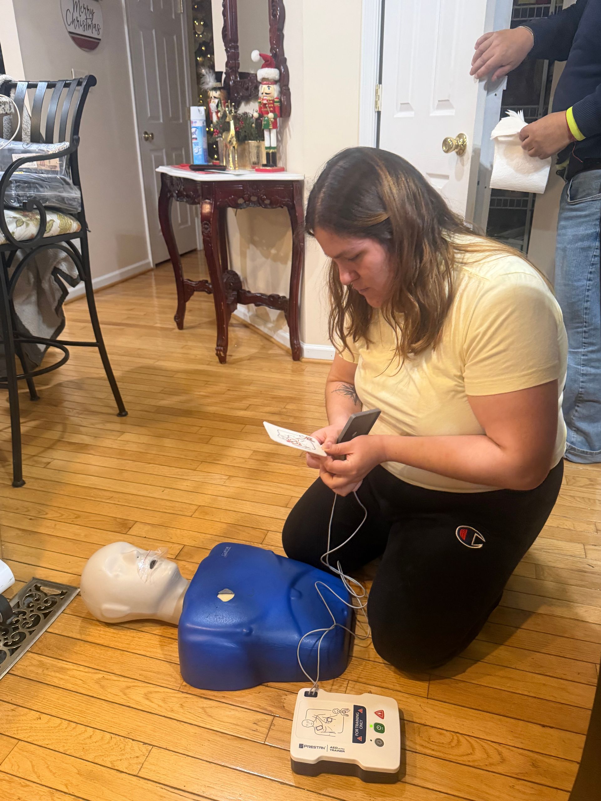 Woman kneeling, practicing CPR on a dummy, with an AED nearby. Inside a room, reading instructions.
