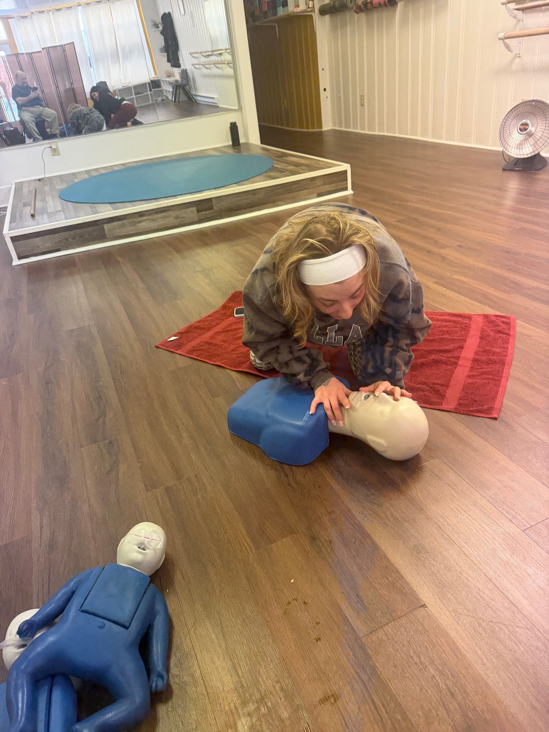 Woman practicing CPR on a training dummy, indoors. Blue and white training equipment on a wooden floor.