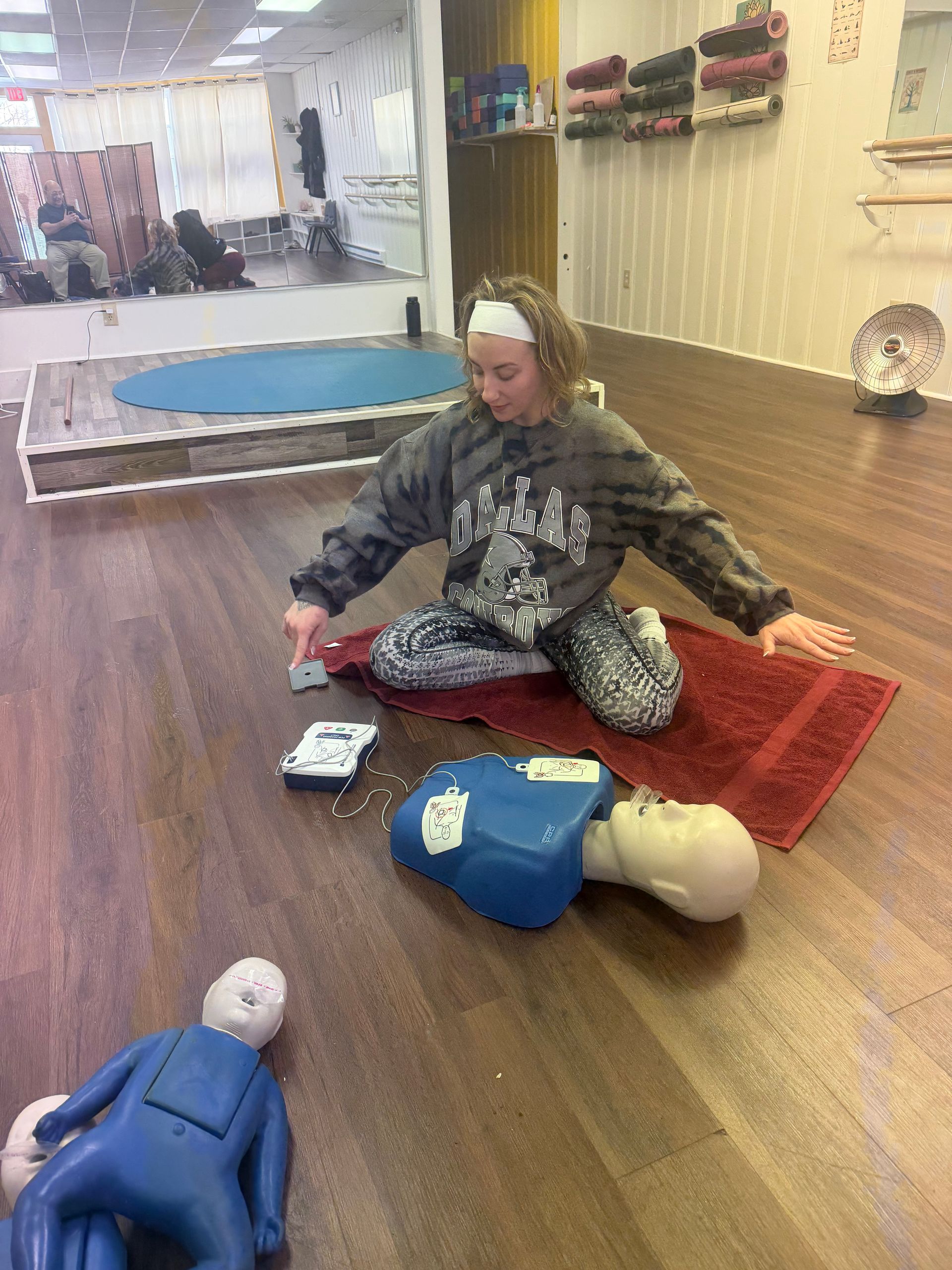 Woman practices CPR on a training dummy, kneeling on a red mat in a dance studio.