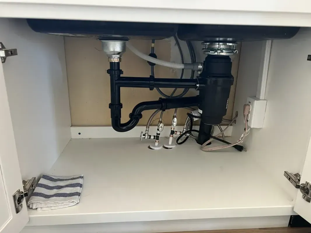Under-sink cabinet with plumbing: black pipes, disposal unit, silver drain pipes, and electrical outlet. White cabinet.