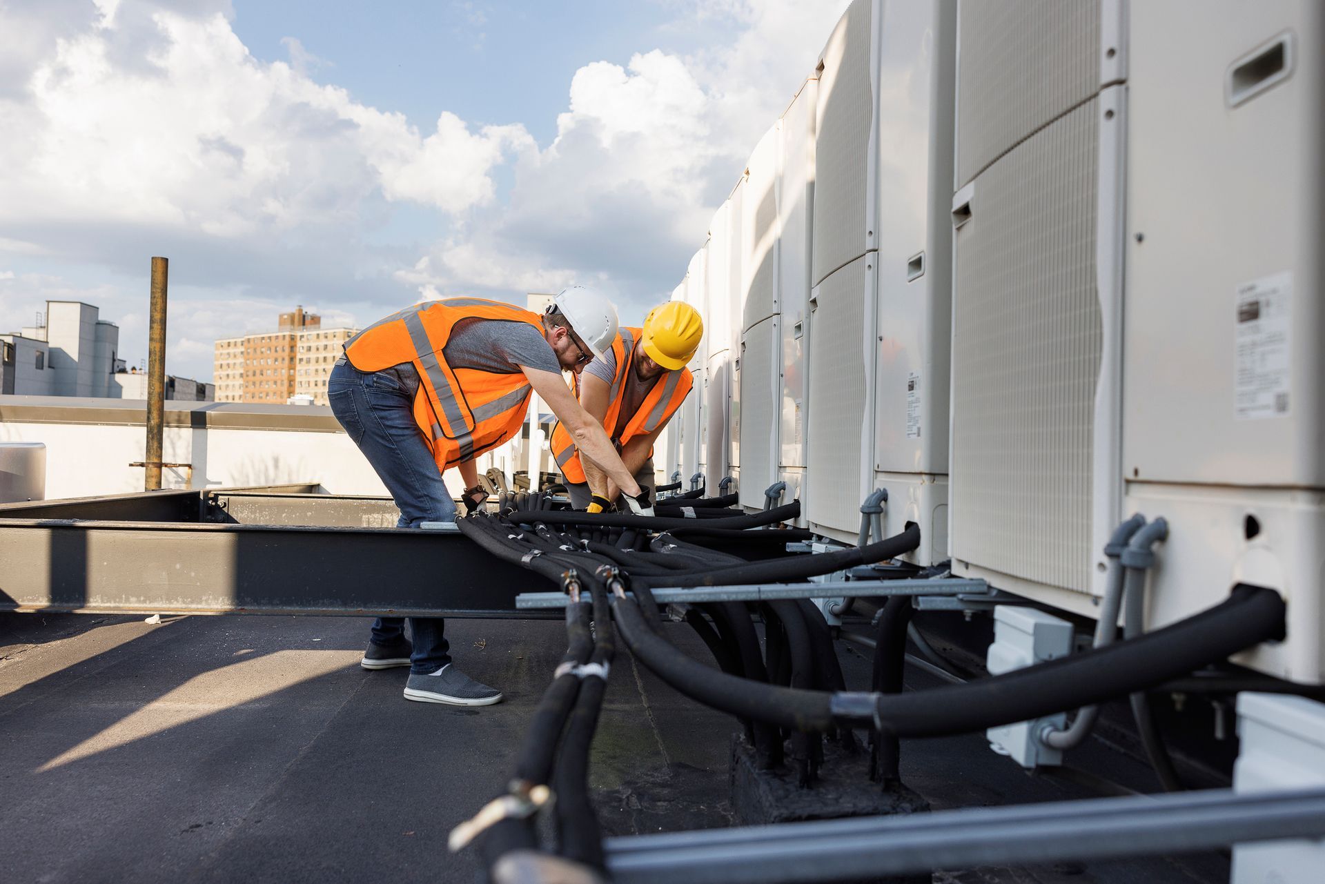 Two workers on a rooftop, wearing safety vests and helmets, working on AC units.