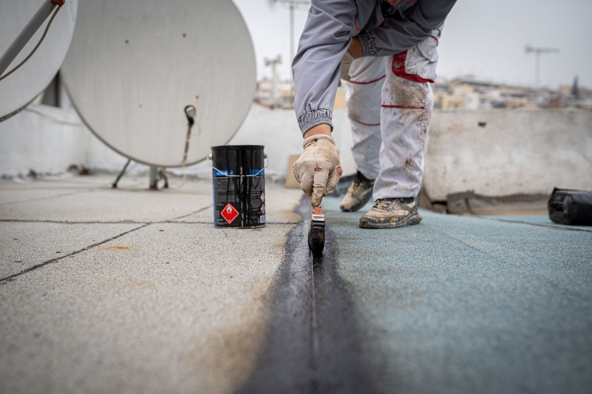 Person in work clothes applying sealant to a roof seam with a brush, alongside a paint can.