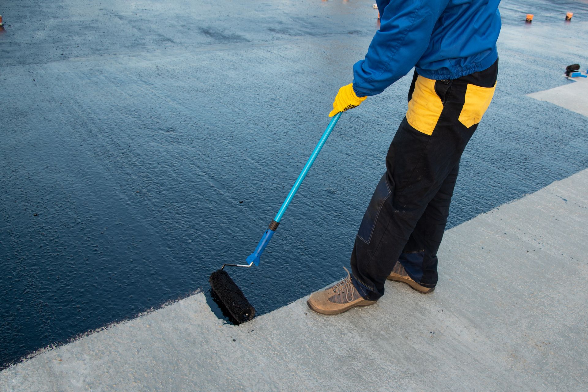 Person in blue jacket and yellow gloves rolling dark sealant on a gray concrete surface.
