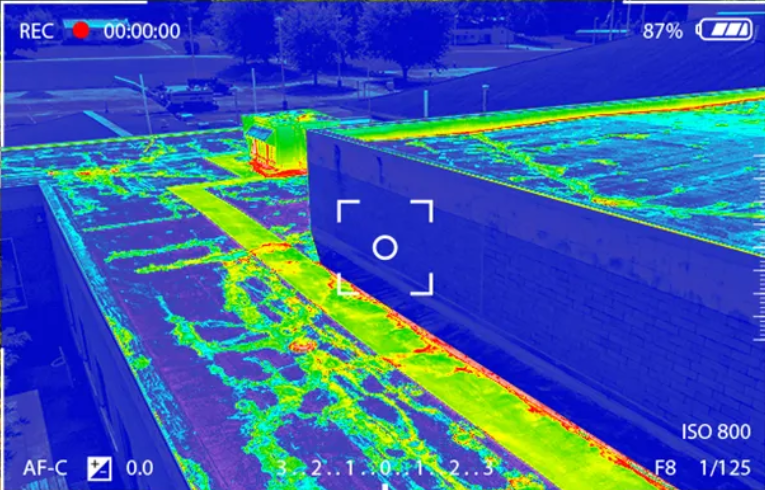 Thermal view of a building roof, showing heat variations in blue, green, and red, with a targeting reticle.