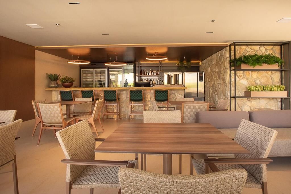 Área de bar interna com mesas, cadeiras, banquetas e uma parede com detalhes em pedra.