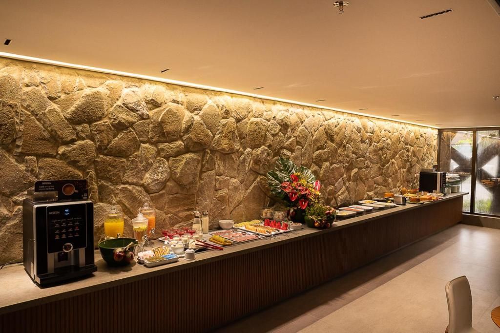 Buffet de café da manhã com máquina de café, suco, doces e flores, disposto junto a uma parede de pedra.