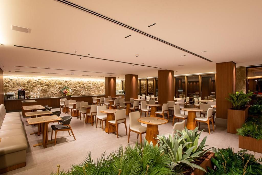 Interior de restaurante com mesas, cadeiras, plantas e iluminação aconchegante.