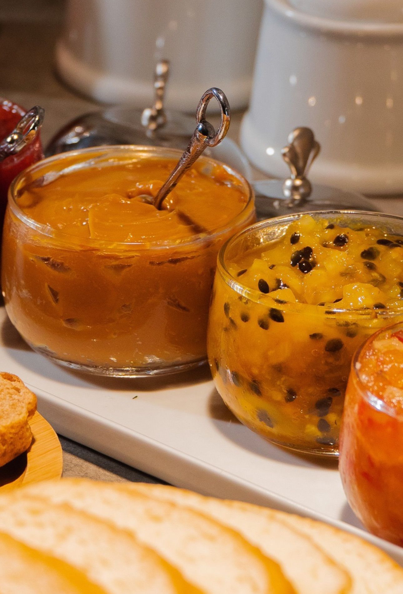 Potes de marmelada, geleia de maracujá e fatias de pão em uma tábua de servir.