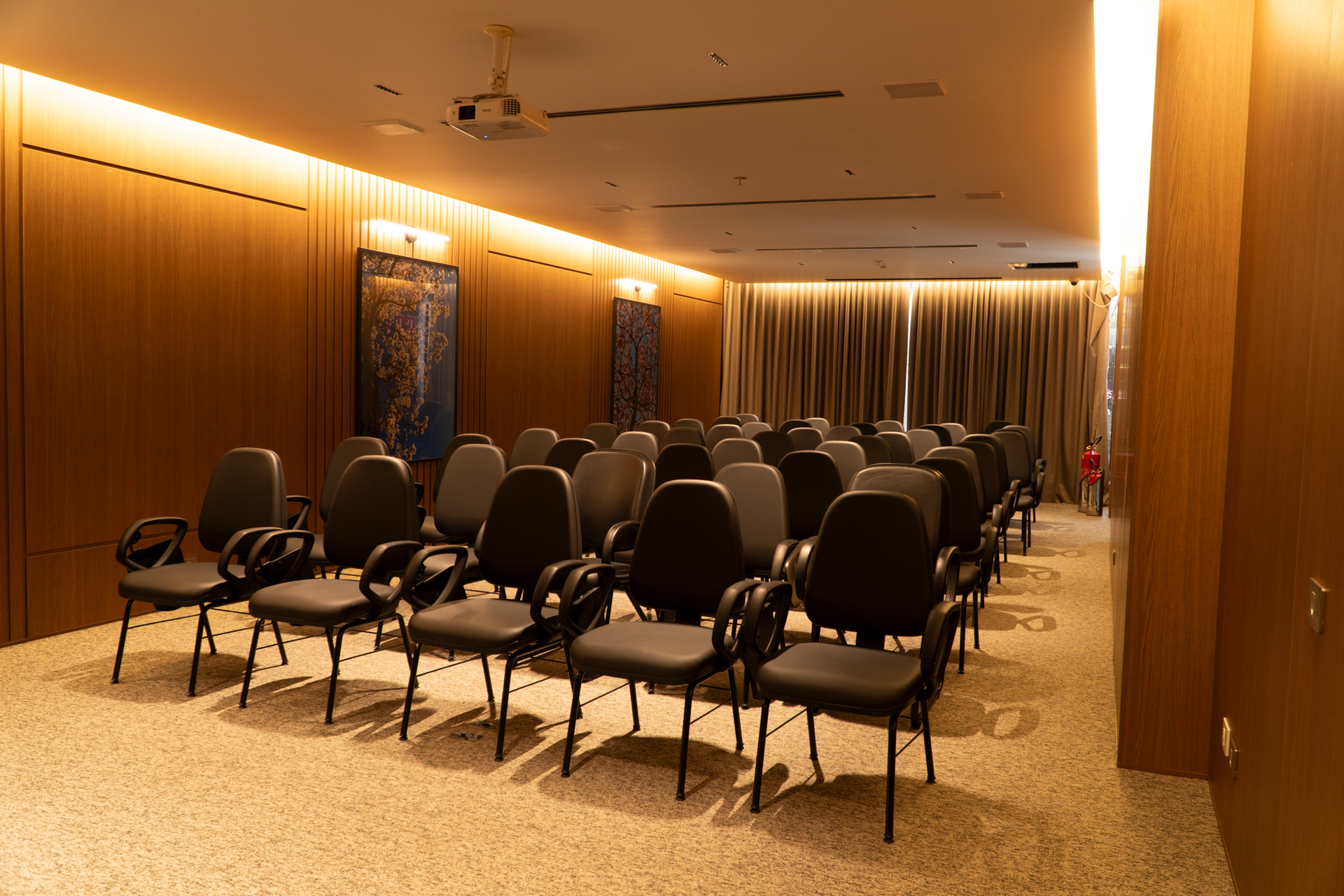Uma sala de apresentações com fileiras de cadeiras pretas, projetor, paredes em tons neutros e obras de arte.