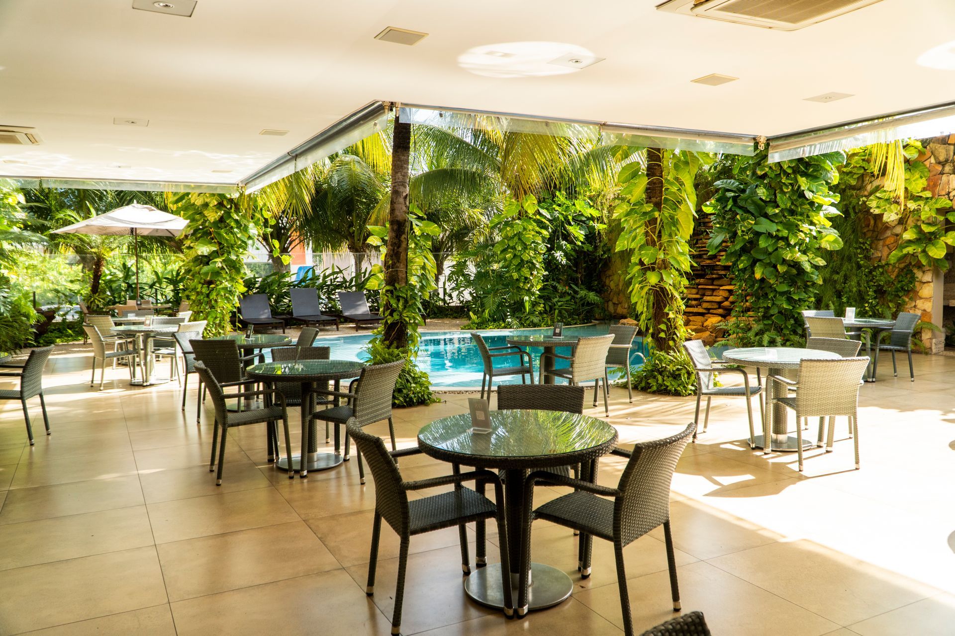 Área de refeições ao ar livre com mesas e cadeiras perto da piscina, rodeada por vegetação exuberante.