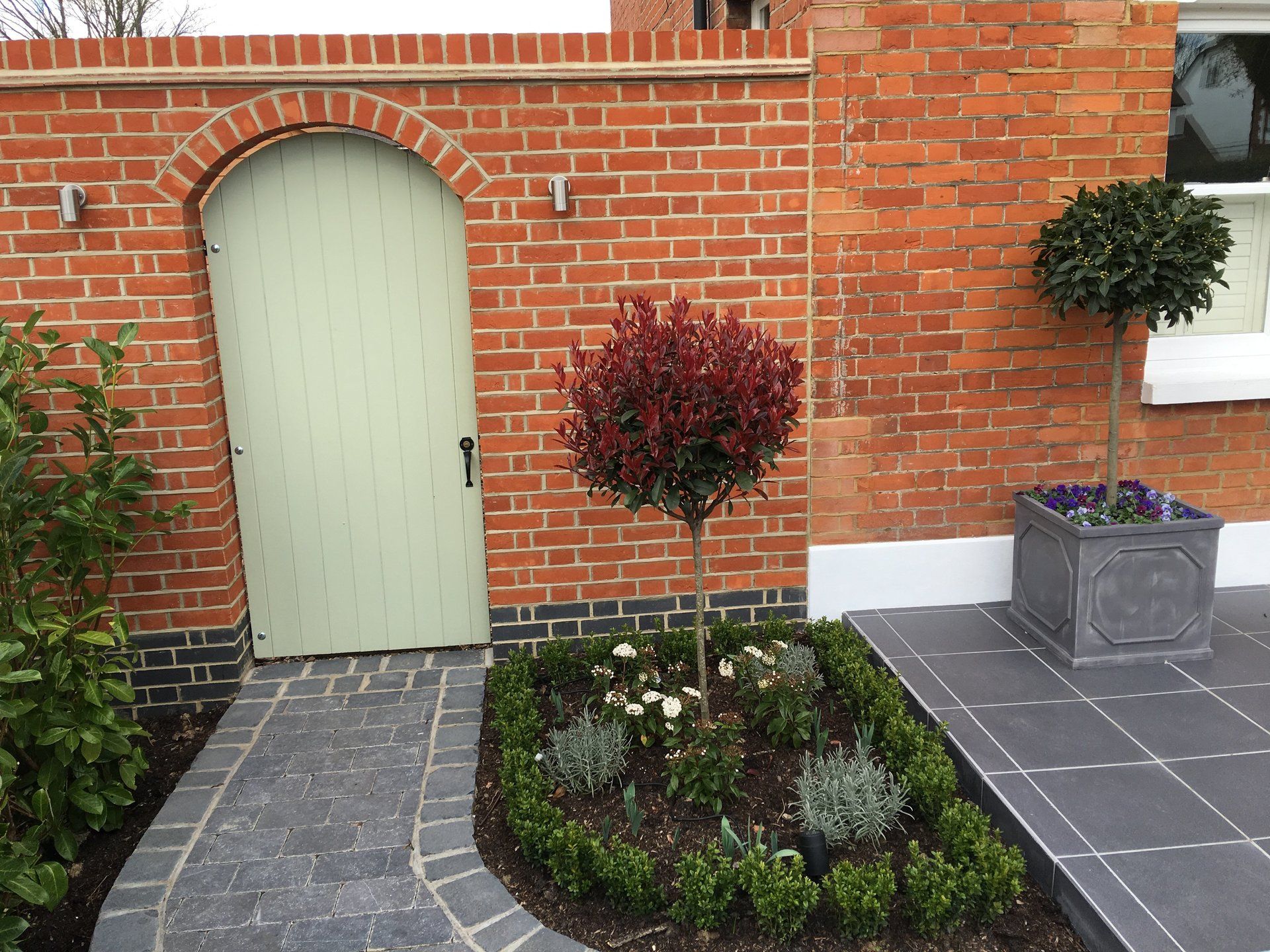 A brick wall with a green door and a garden in front of it.