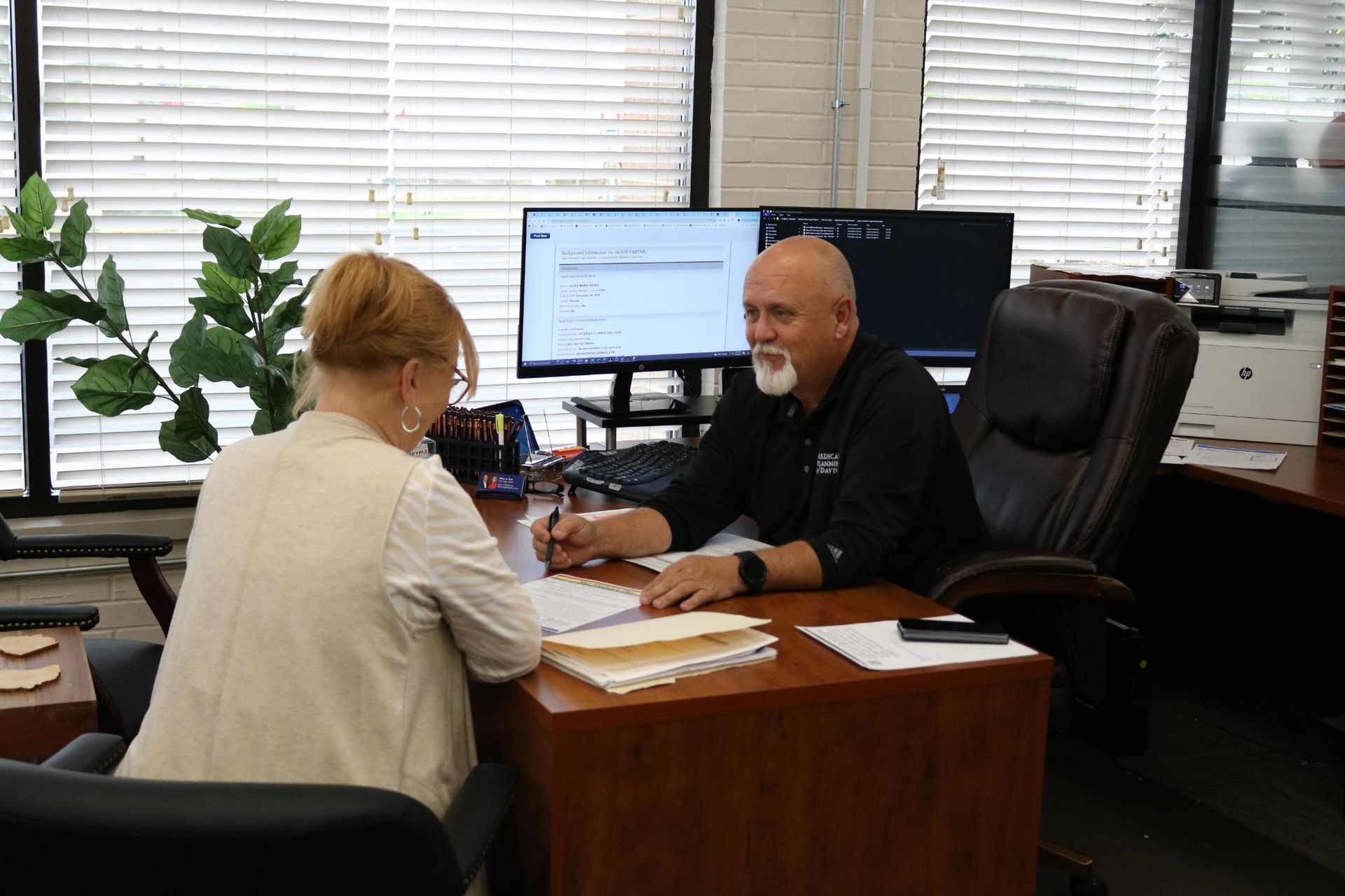 Chris at desk, pen in hand, client across from him, MPD website on monitor.