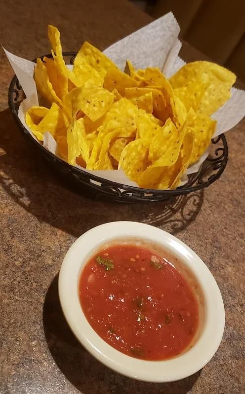 A basket of yellow corn tortilla chips served with a small bowl of red salsa on a speckled table.