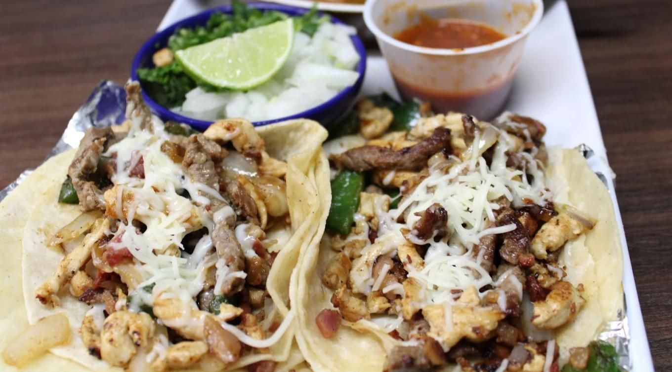Two tacos topped with grilled meat and melted cheese, served with sides of diced onions, cilantro, lime, and red salsa.